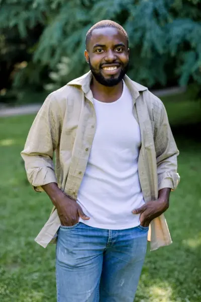 Summer outdoor portrait of handsome smiling African man in jeans white t shirt and beige casual shirt looking at camera while standing on green grass in park enjoying the walk